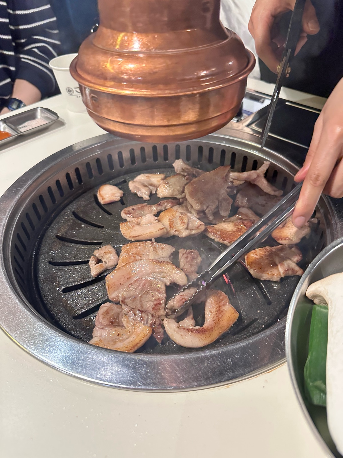 A staff member grilling aged Jeju pork with tongs under a copper smoke hood at Gojip132